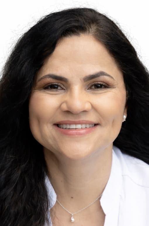 Smiling woman with long dark wavy hair, wearing a white shirt and pearl necklace.